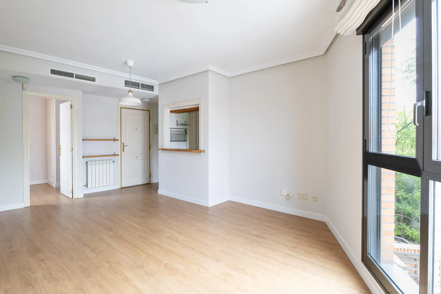 Bright, empty room with light wood floors, white walls, and a large window with a view of trees.