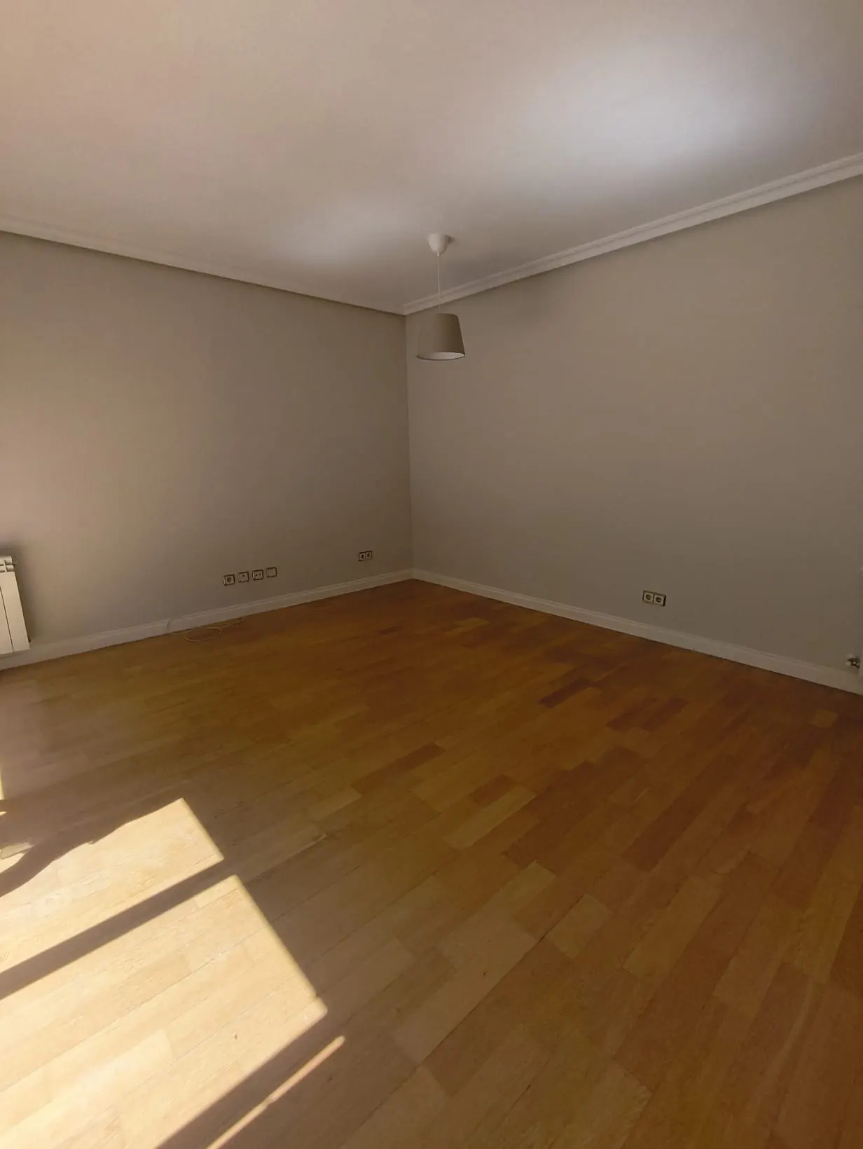 Empty room with light beige walls, polished hardwood floors, and a single pendant light fixture hanging from the ceiling.