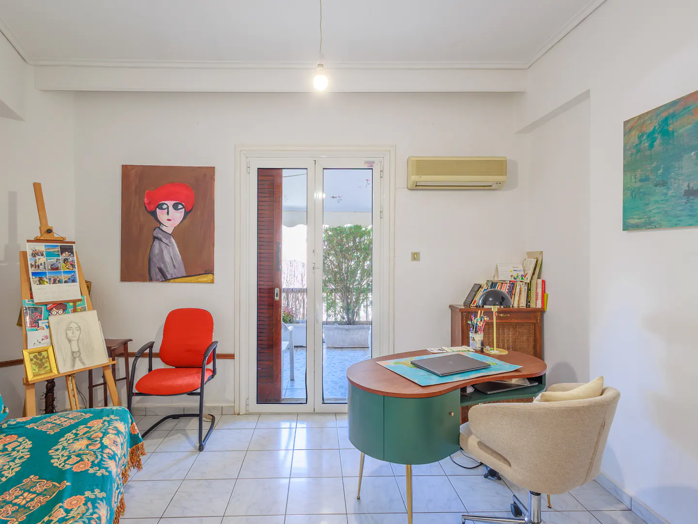 Bright room with white walls, tiled floor, and art. A green desk, beige chair, and red chair are present. Paintings and an easel add to the artistic vibe.