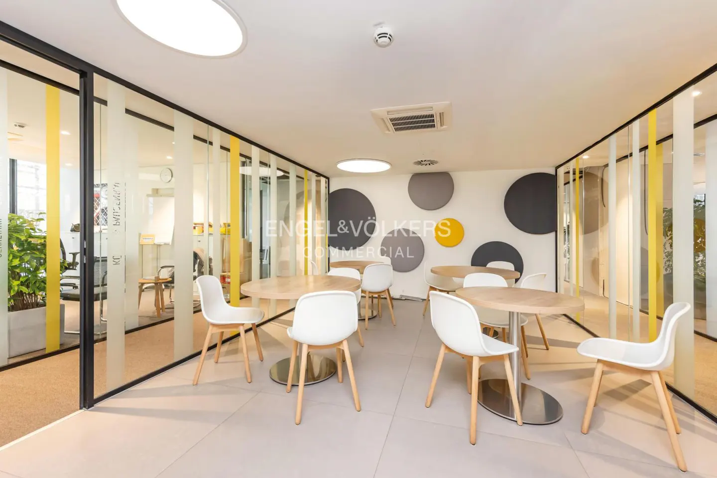 Bright office space with round tables and white chairs. Engel & Völkers logo on the wall with gray and yellow circles. Glass partitions with yellow stripes.