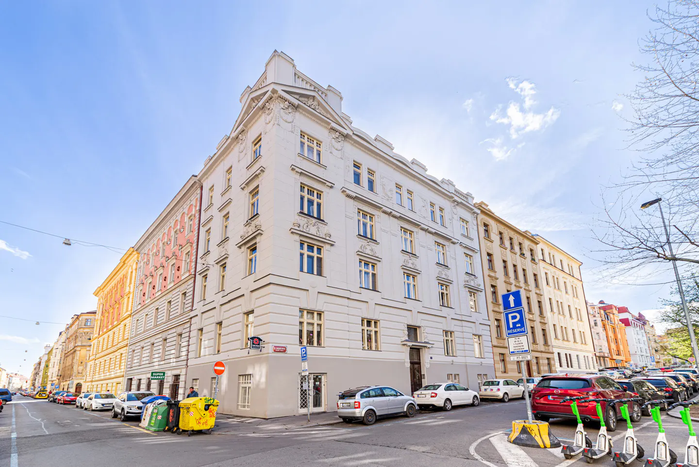 Street view of a light-colored, multi-story building with many windows. Cars and scooters are parked along the street.