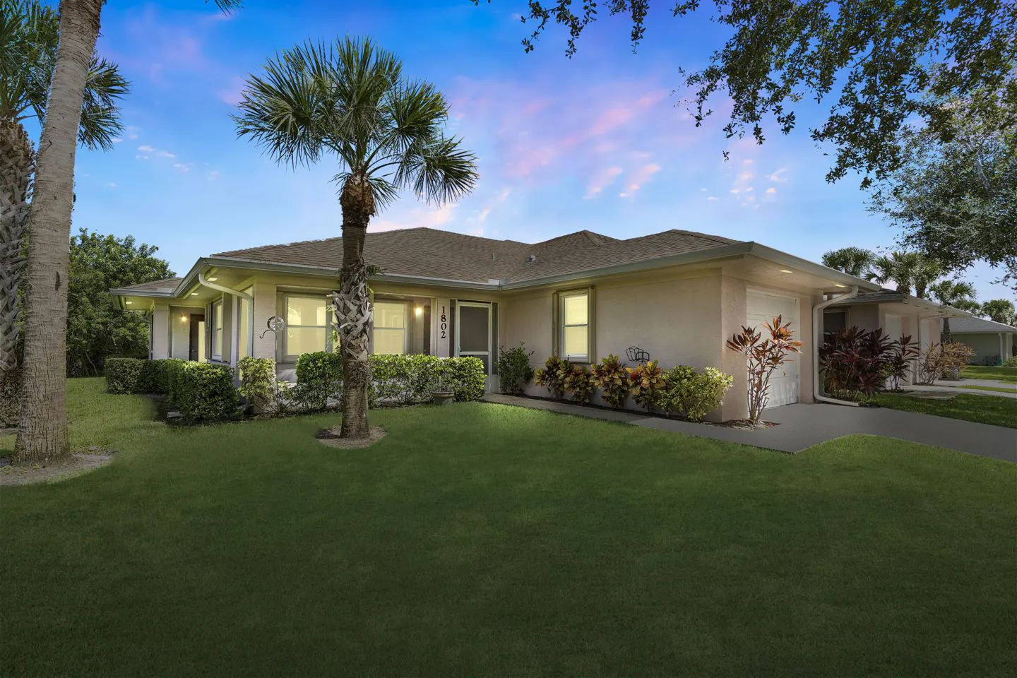 Single-story beige house with a brown roof, palm trees, and a green lawn under a sunset sky.