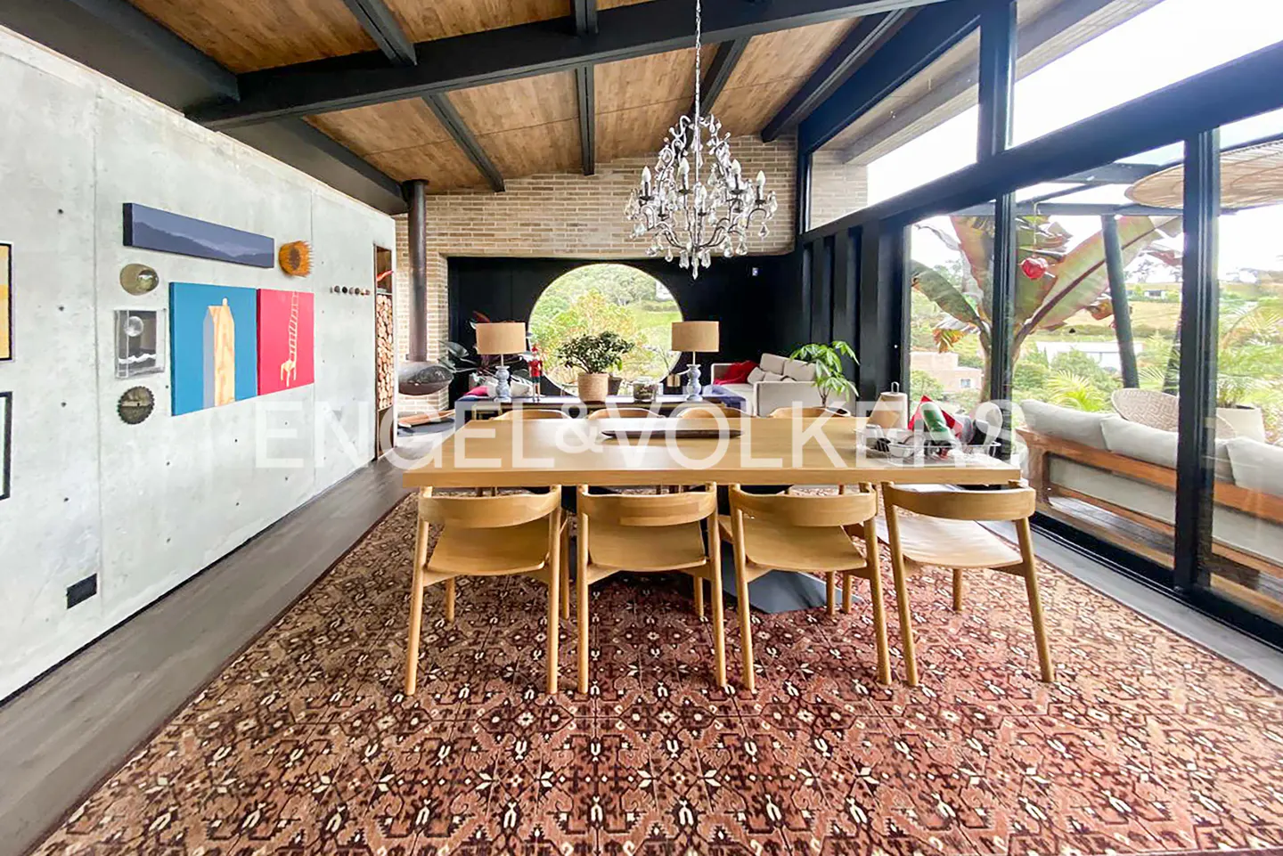 A modern dining room with a wooden table, chairs, and a patterned rug. A chandelier hangs above, and large windows offer a view of greenery.