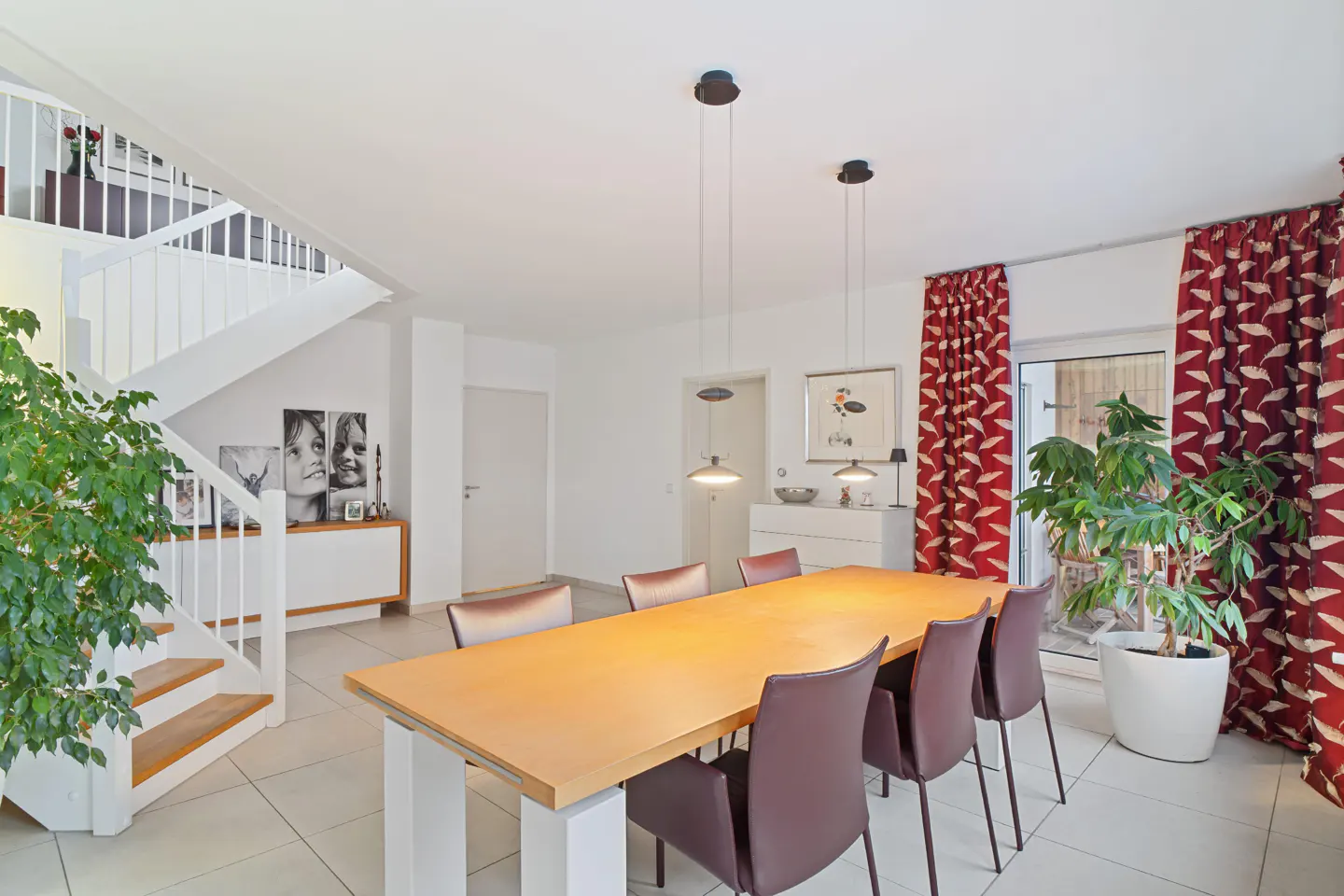 Bright dining room with a long wooden table, six brown chairs, and three pendant lights. Red patterned curtains and a staircase are visible.