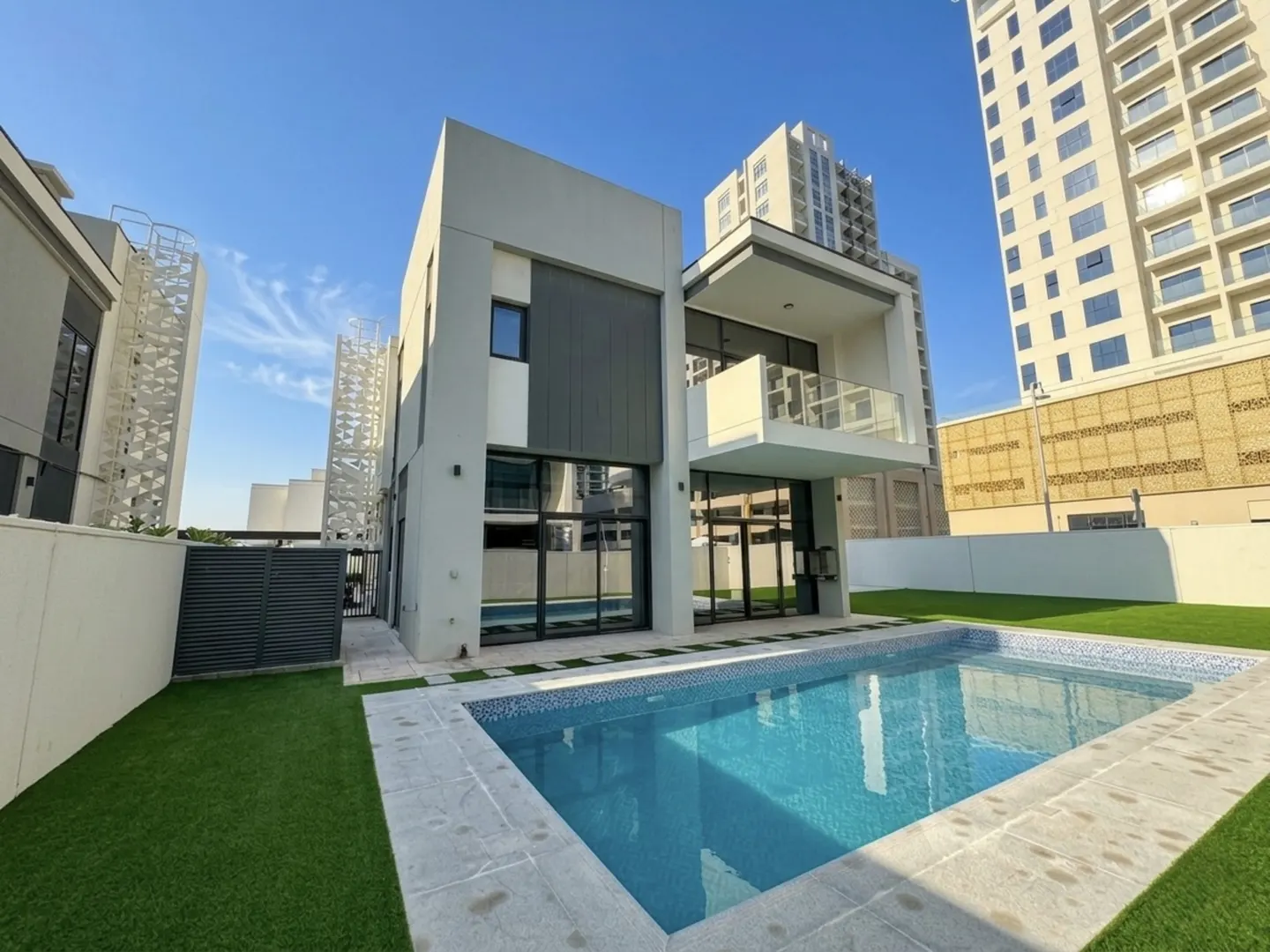 Modern two-story house with a blue tiled swimming pool and green lawn. Tall buildings are in the background.