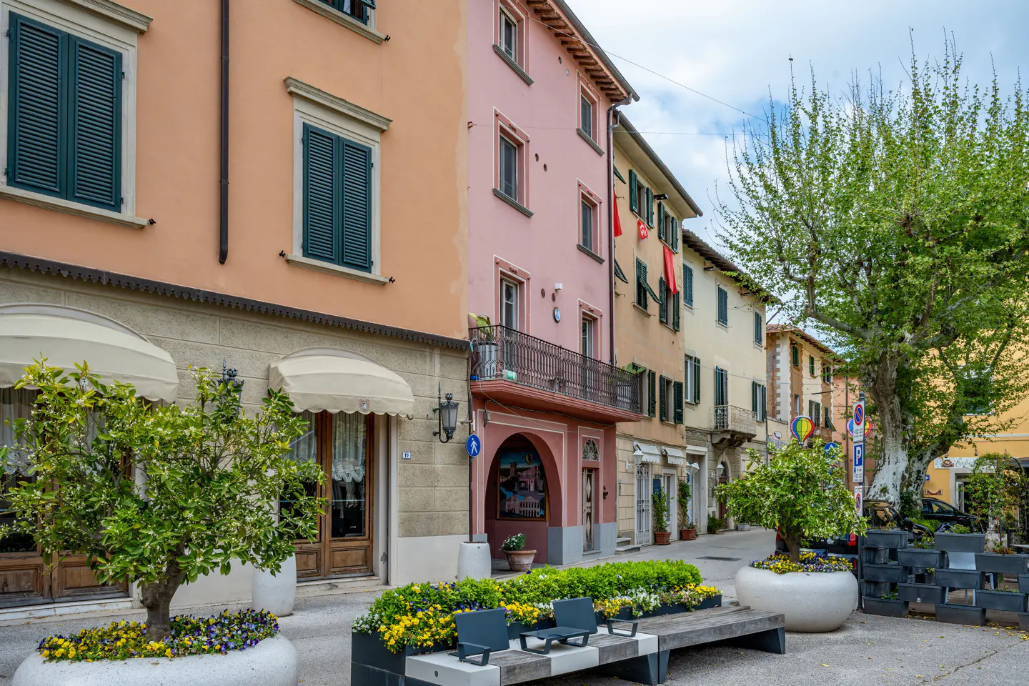 Street view of colorful buildings in Italy. Trees and flowers line the street with a bench in the foreground.