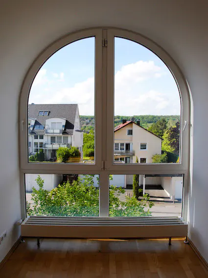 Arched window view of houses and trees under a blue sky. A radiator sits below the window in a room with wood floors.