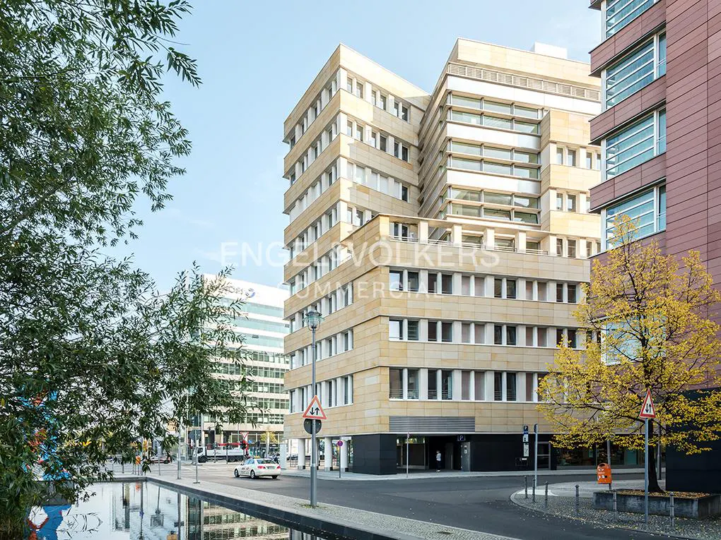 Modern beige office building with white windows, next to a canal and a tree with yellow leaves.