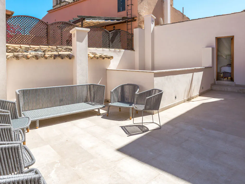 Outdoor patio with gray woven furniture, including a sofa and chairs, on a light stone floor. A building with a pink facade is in the background.