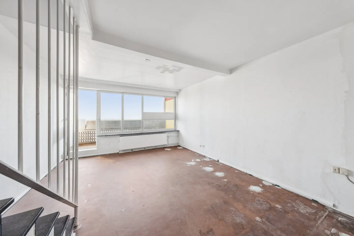 Empty room with white walls, damaged brown floor, and large windows. A metal staircase is on the left.