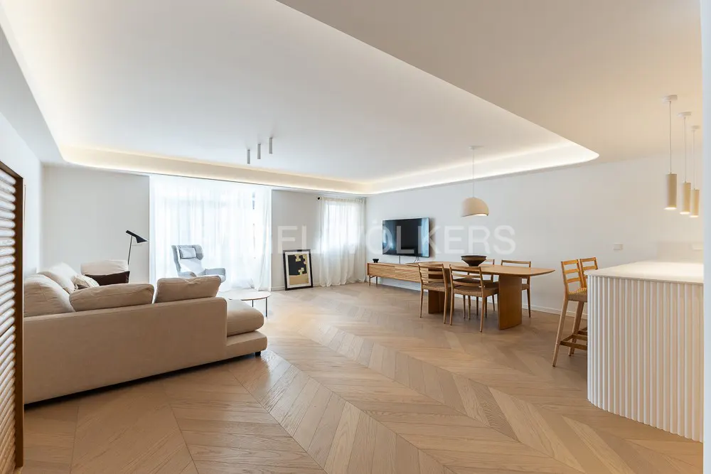 Bright, open-concept living space with herringbone wood floors. Beige sofa, dining table, and kitchen island. Natural light from large windows.