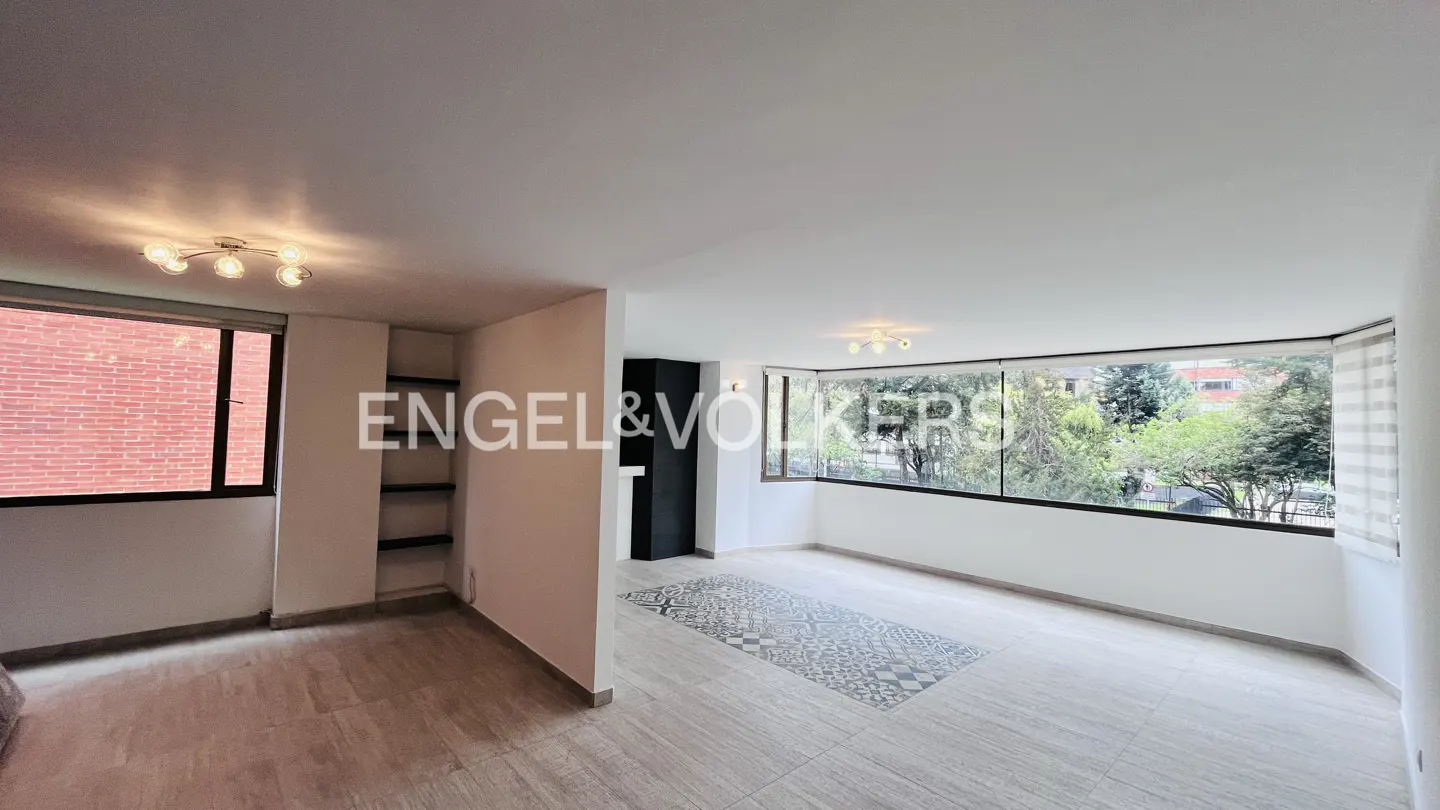 Bright, empty apartment with large windows overlooking trees. The floor is tiled and there is a decorative rug. Engel & Volkers logo is visible.