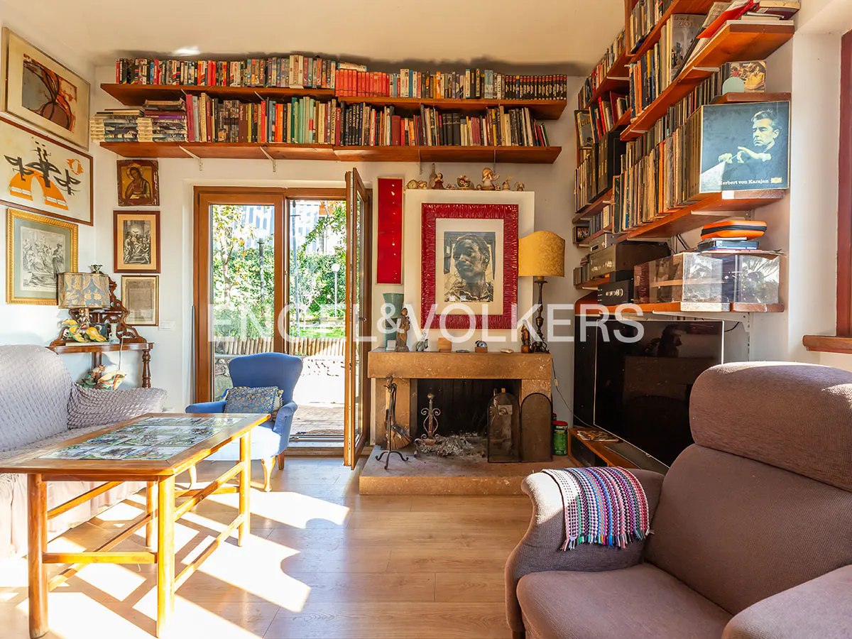 Cozy living room with bookshelves, a fireplace, two armchairs, a wooden coffee table, and glass doors leading to a garden.