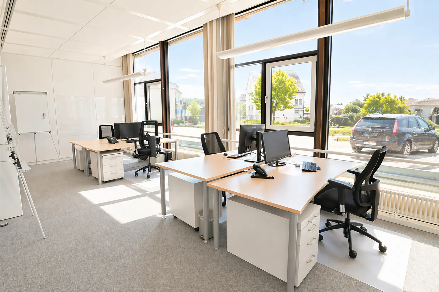 Bright office space with desks, computers, and black chairs. Large windows offer a view of a car and greenery outside.