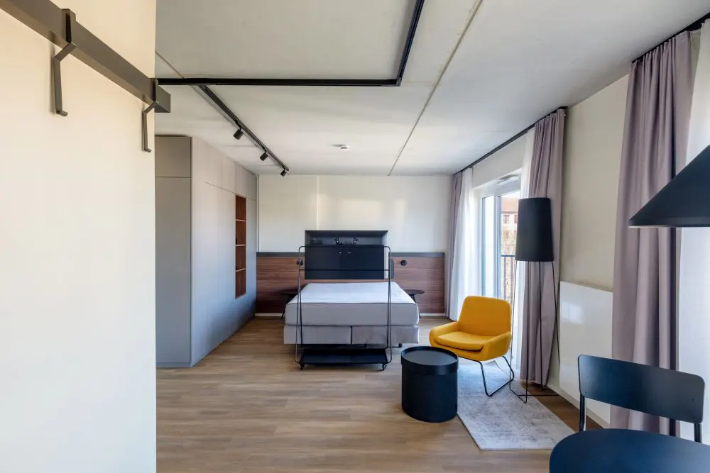 A modern bedroom with a bed, yellow chair, black table, and gray curtains. The room has wood floors and track lighting.