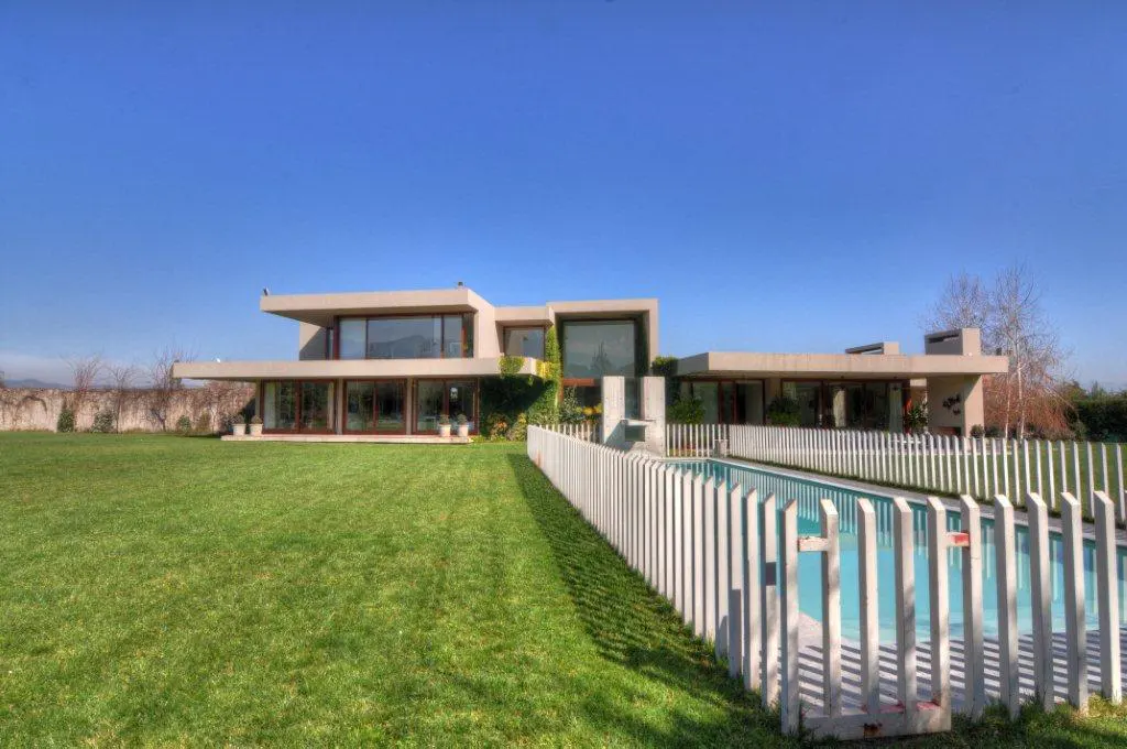 Modern beige house with large windows, green lawn, and a pool with a white fence under a blue sky.