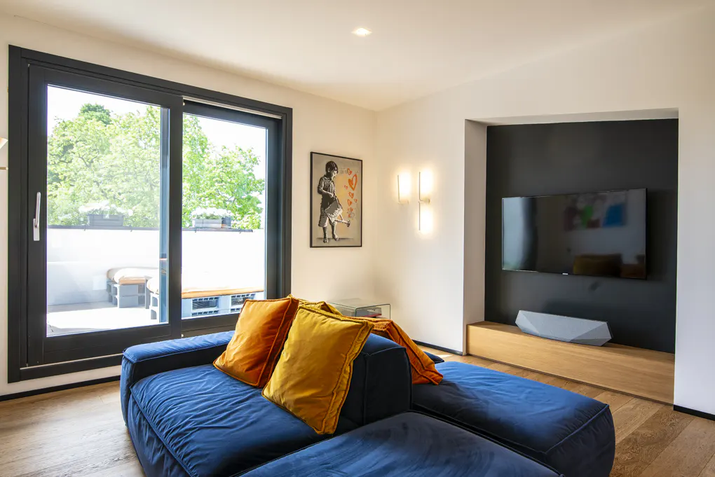 Living room with a blue sofa, orange pillows, a TV, and a balcony with a view of trees.