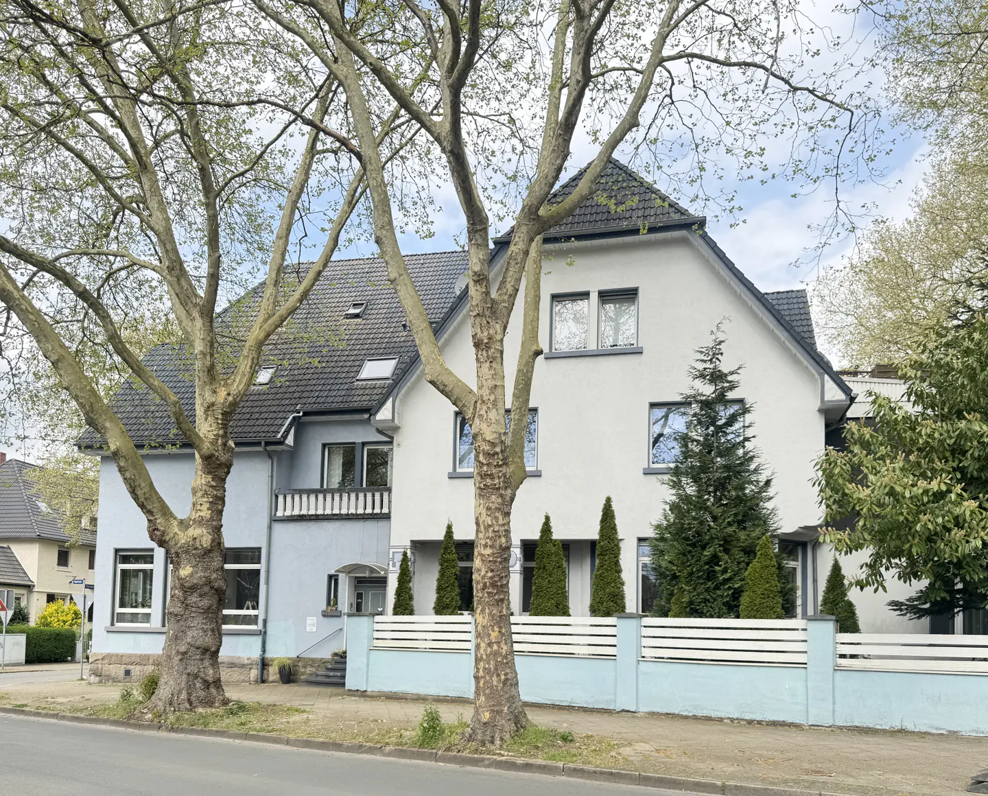 Two-story house with a gray roof and white and light blue exterior, framed by trees and a white fence.