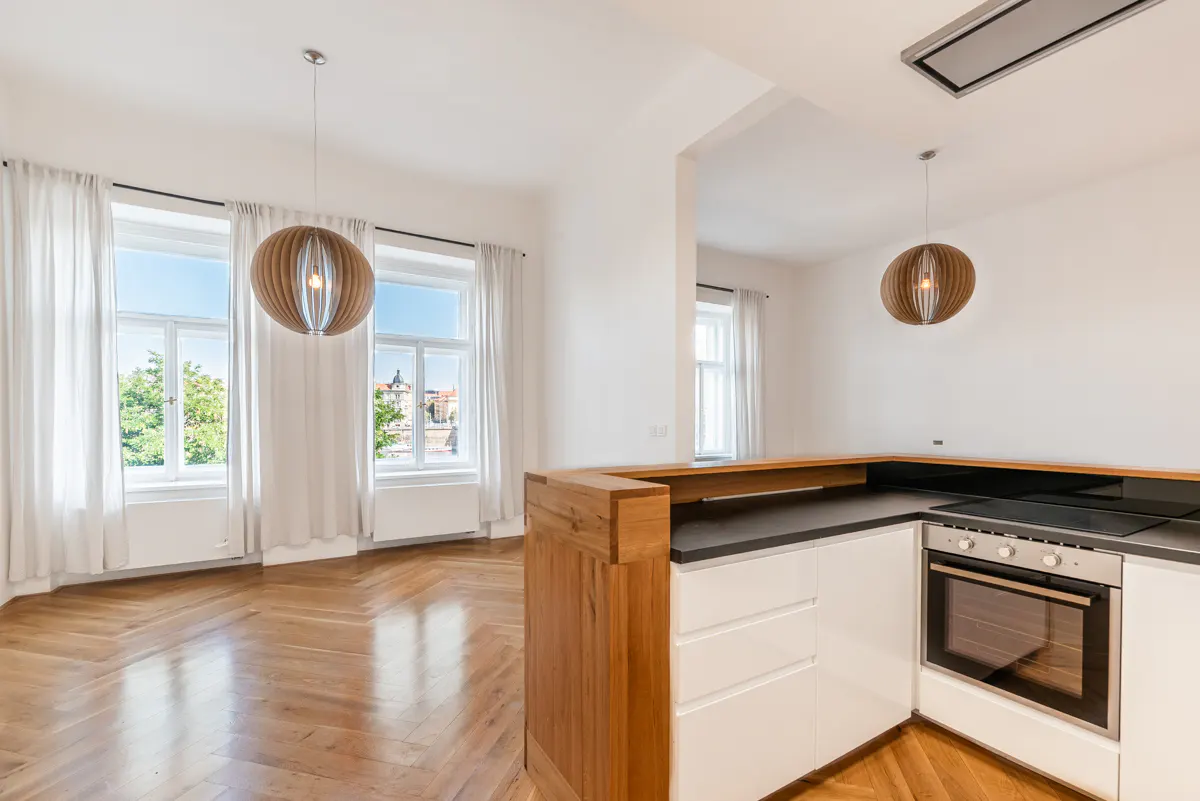 Bright, modern kitchen with white cabinets, black countertops, and stainless steel oven. Large windows with white curtains overlook a cityscape. Wooden floors and pendant lights.