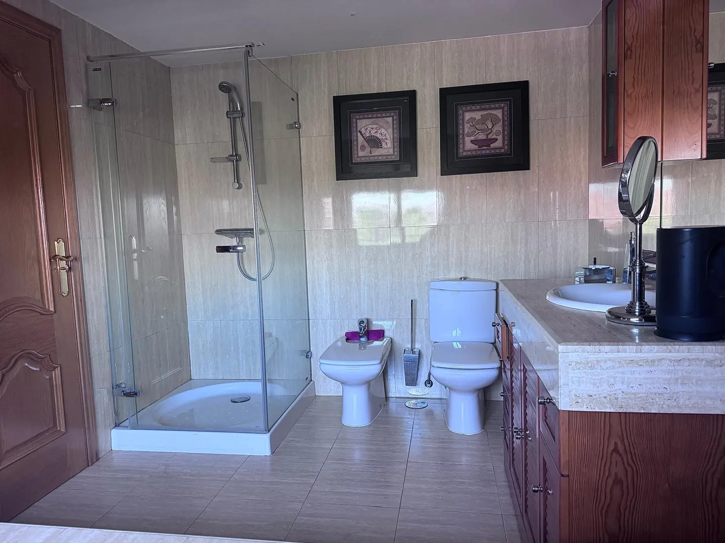 Bathroom with a glass shower, toilet, bidet, and vanity with a sink and mirror. Two framed pictures hang on the tiled wall.