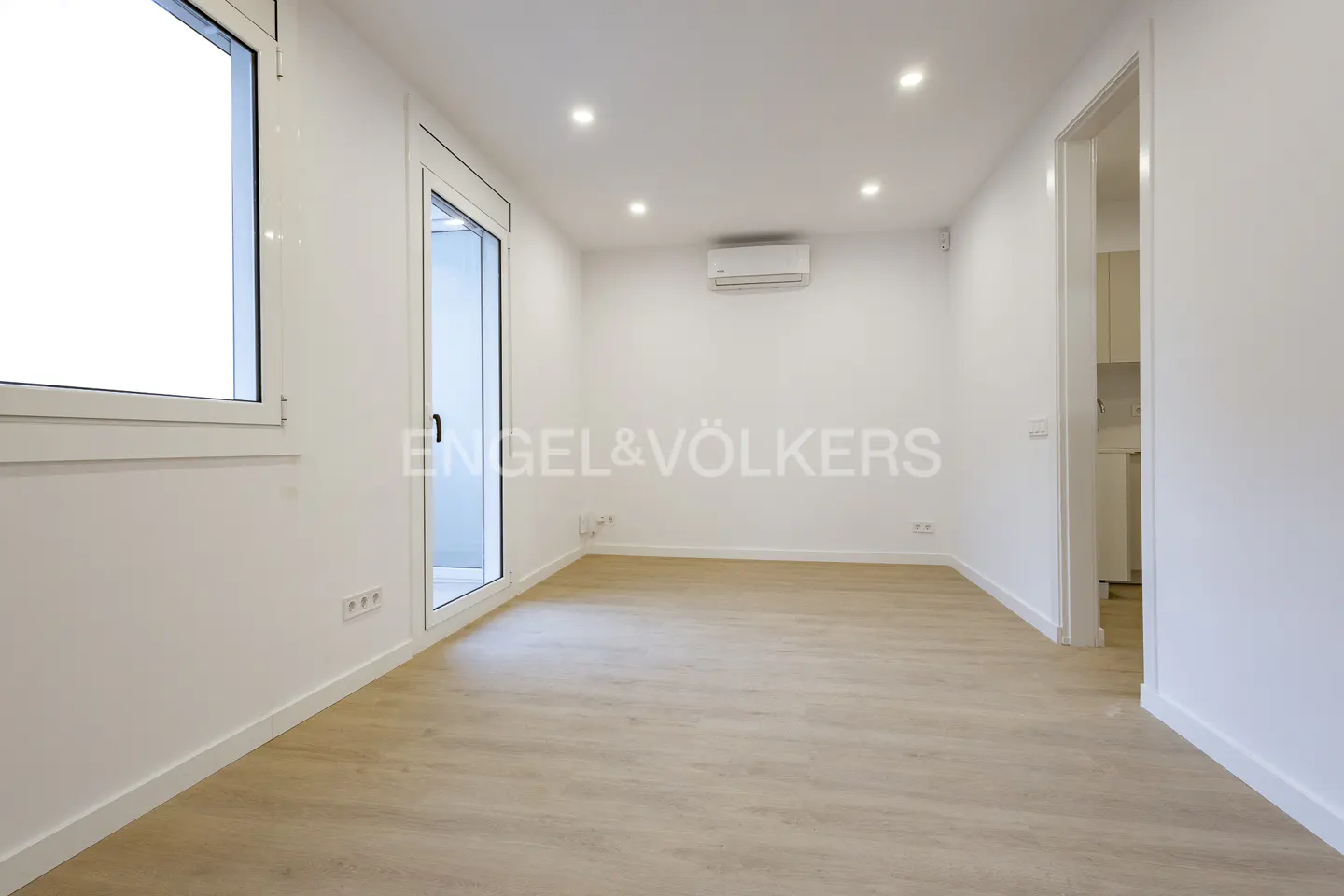 Bright, empty room with light wood floors, white walls, and recessed lighting. A window and door let in natural light. Engel & Völkers logo on the wall.