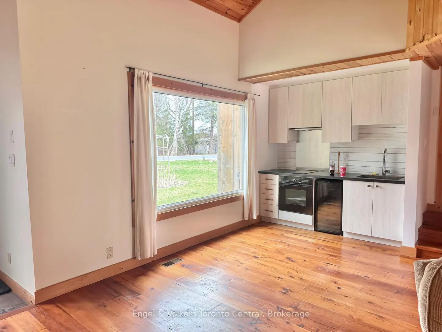 Interior view of a room with hardwood floors, a large window with white curtains, and a small kitchen area with light wood cabinets.