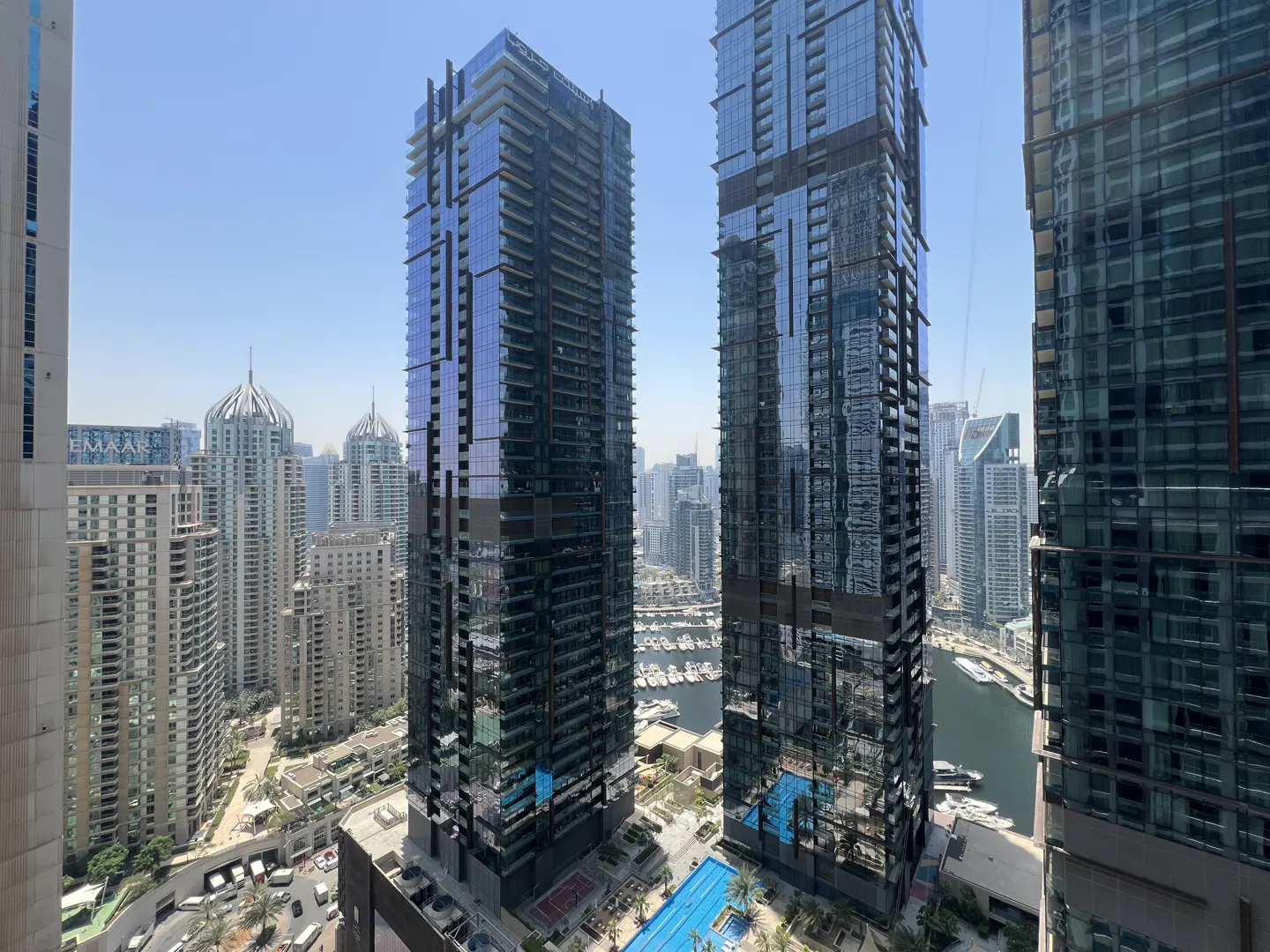 View of Dubai Marina with tall glass skyscrapers, a blue pool, and boats on the water under a clear sky.