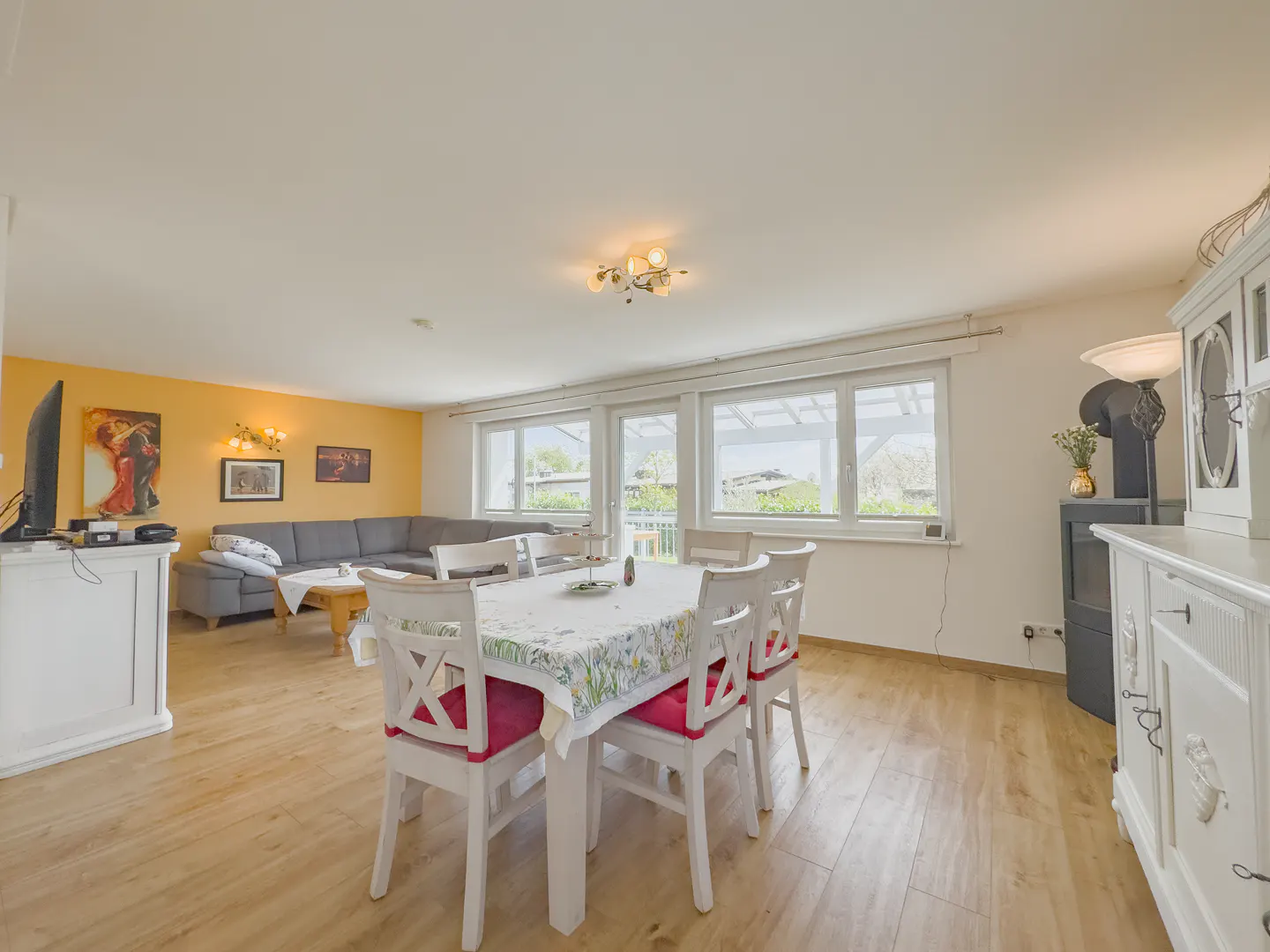 Bright, open-concept living and dining area with wood floors. White dining table with floral tablecloth and red-cushioned chairs. Gray sofa and yellow accent wall.