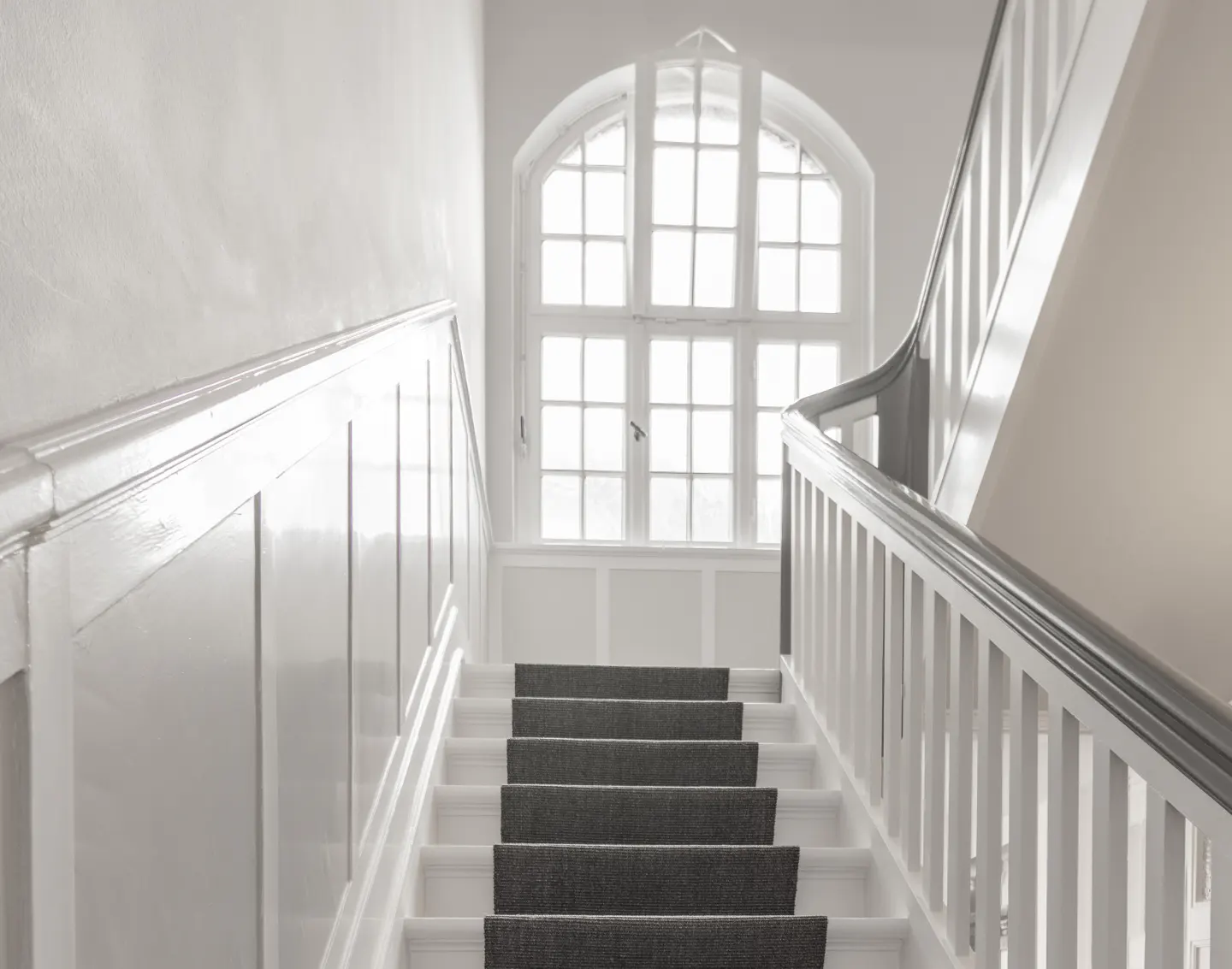 White staircase with gray carpet runner leading to a large arched window.