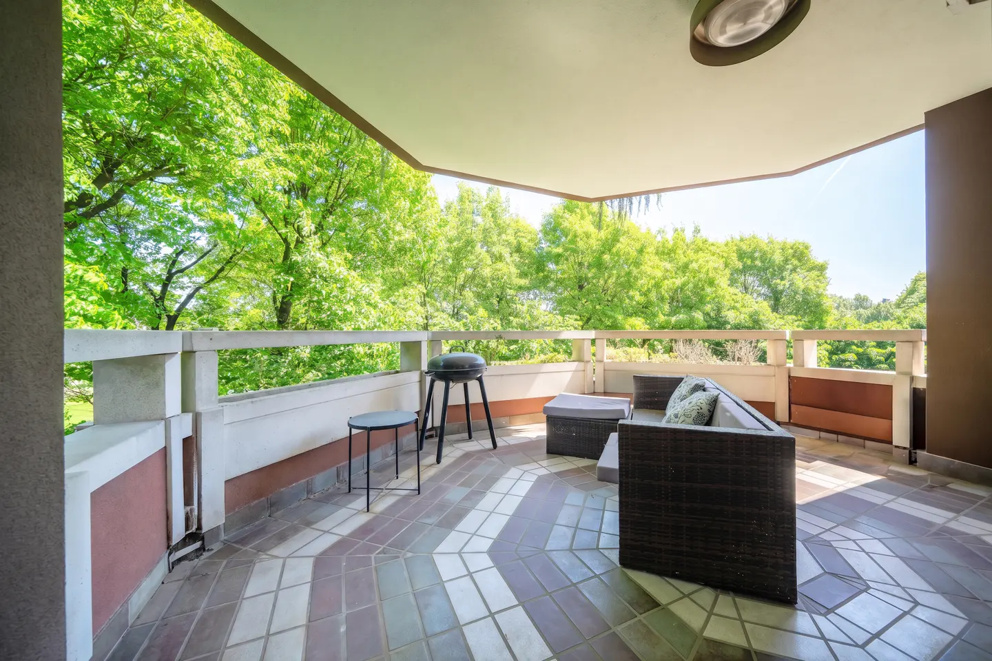 A covered balcony with a grill, table, and wicker sofa overlooks lush green trees. The floor is tiled in a geometric pattern.