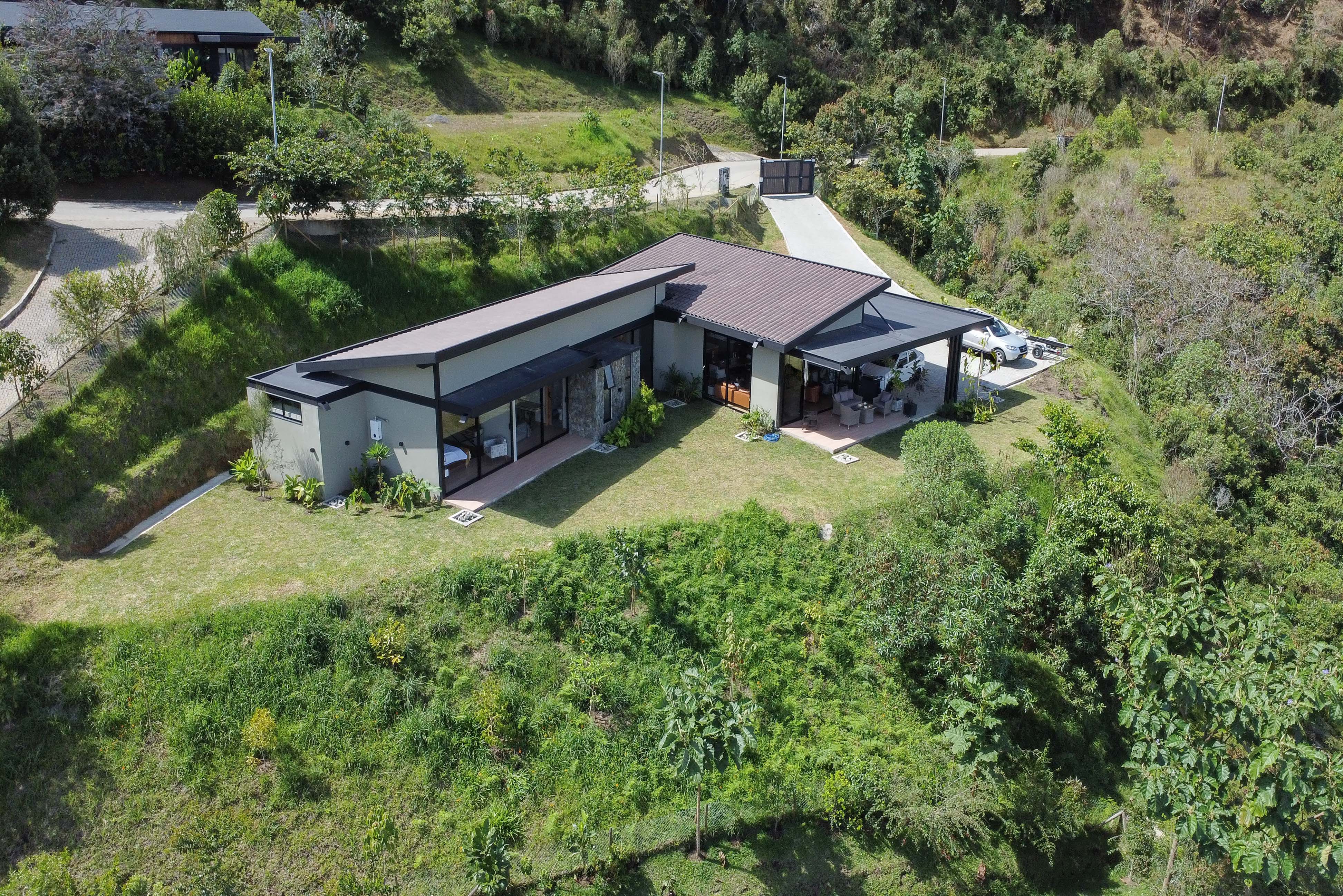 Aerial view of a modern, light-colored home with a dark roof, nestled in a lush, green, hilly landscape.