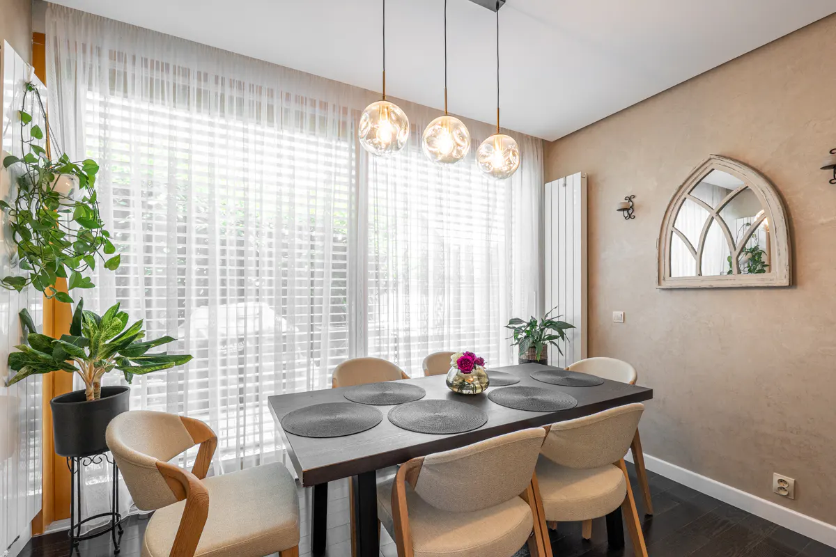 Dining room with a dark wood table, six beige chairs, and three pendant lights. A large window with sheer curtains is on the left.