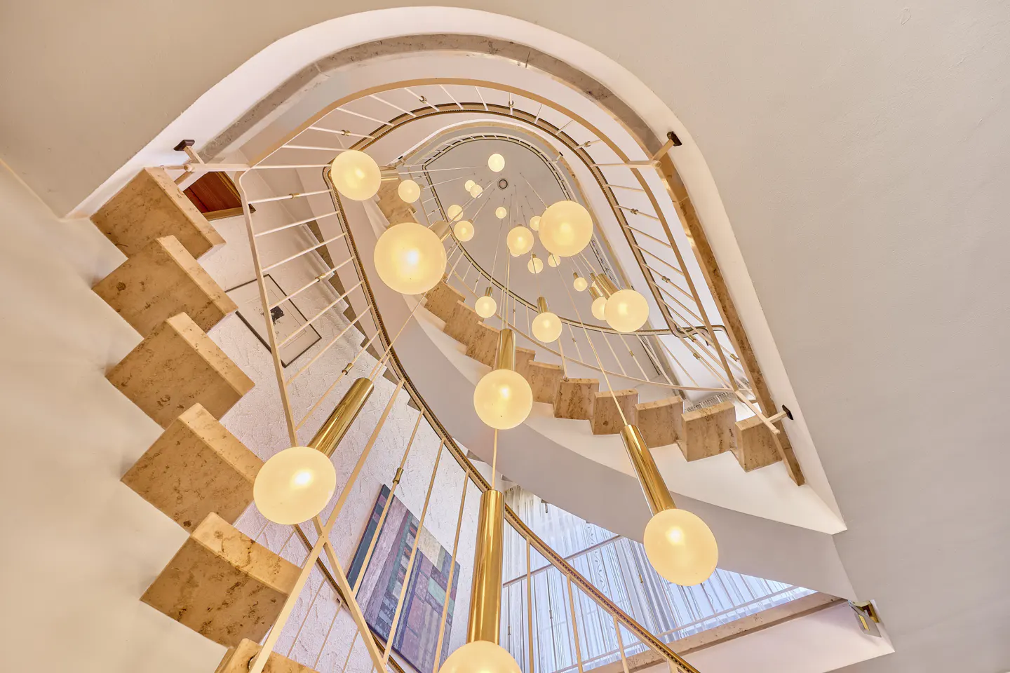 Looking up at a spiral staircase with stone steps, white railings, and a modern chandelier with globe lights.