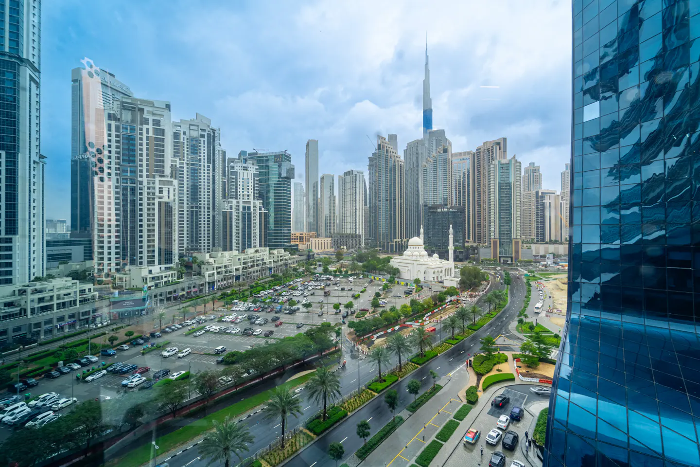 Dubai skyline view featuring skyscrapers, a white mosque, and a parking lot, under a cloudy sky.