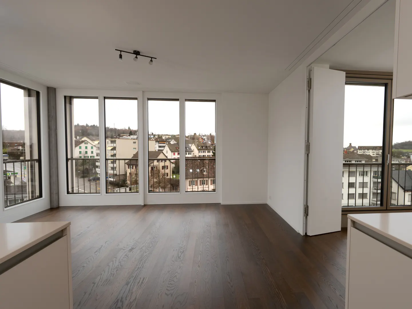 Bright, empty room with dark wood floors and white walls. Large windows offer a city view. A modern light fixture is on the ceiling.