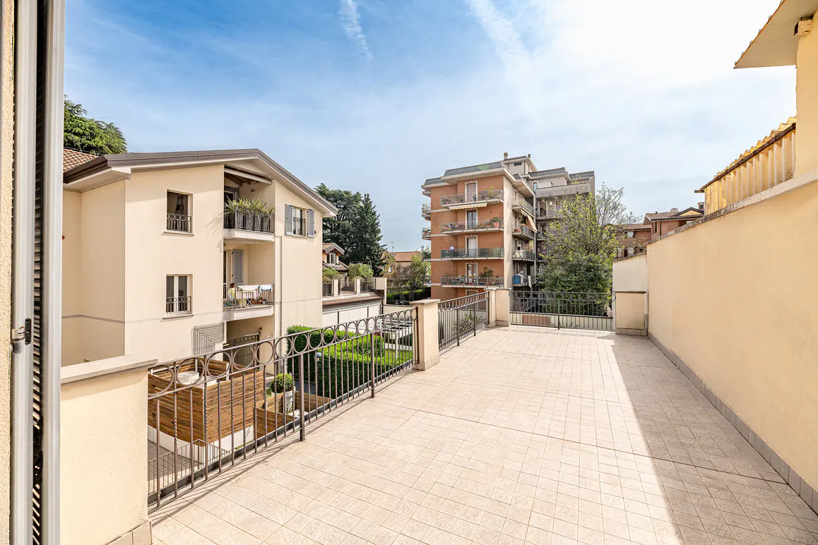View from a tiled balcony with metal railings, overlooking a garden and buildings under a blue sky.