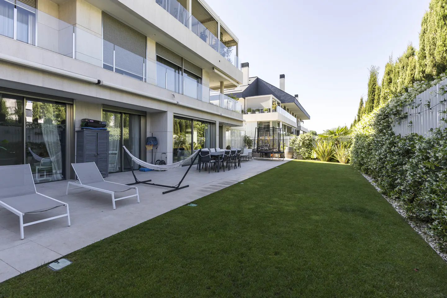 Backyard view of a modern, multi-story home with a green lawn, patio furniture, hammock, and a trampoline in the background.
