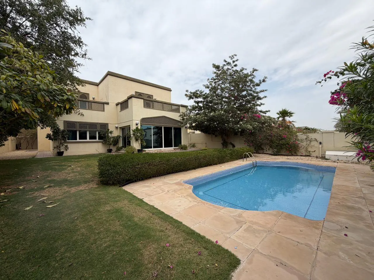 Two-story beige house with a blue swimming pool, green grass, trees, and a stone patio on a cloudy day.