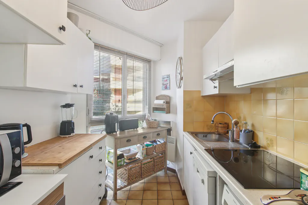 A bright kitchen with white cabinets, a wood countertop, and a window with blinds. A wooden table with baskets sits near the window.