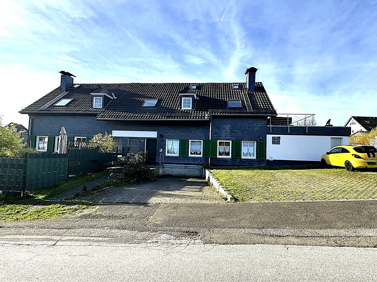 Two-story house with dark gray siding, green shutters, and a black roof. A yellow car is parked on the right side of the house.