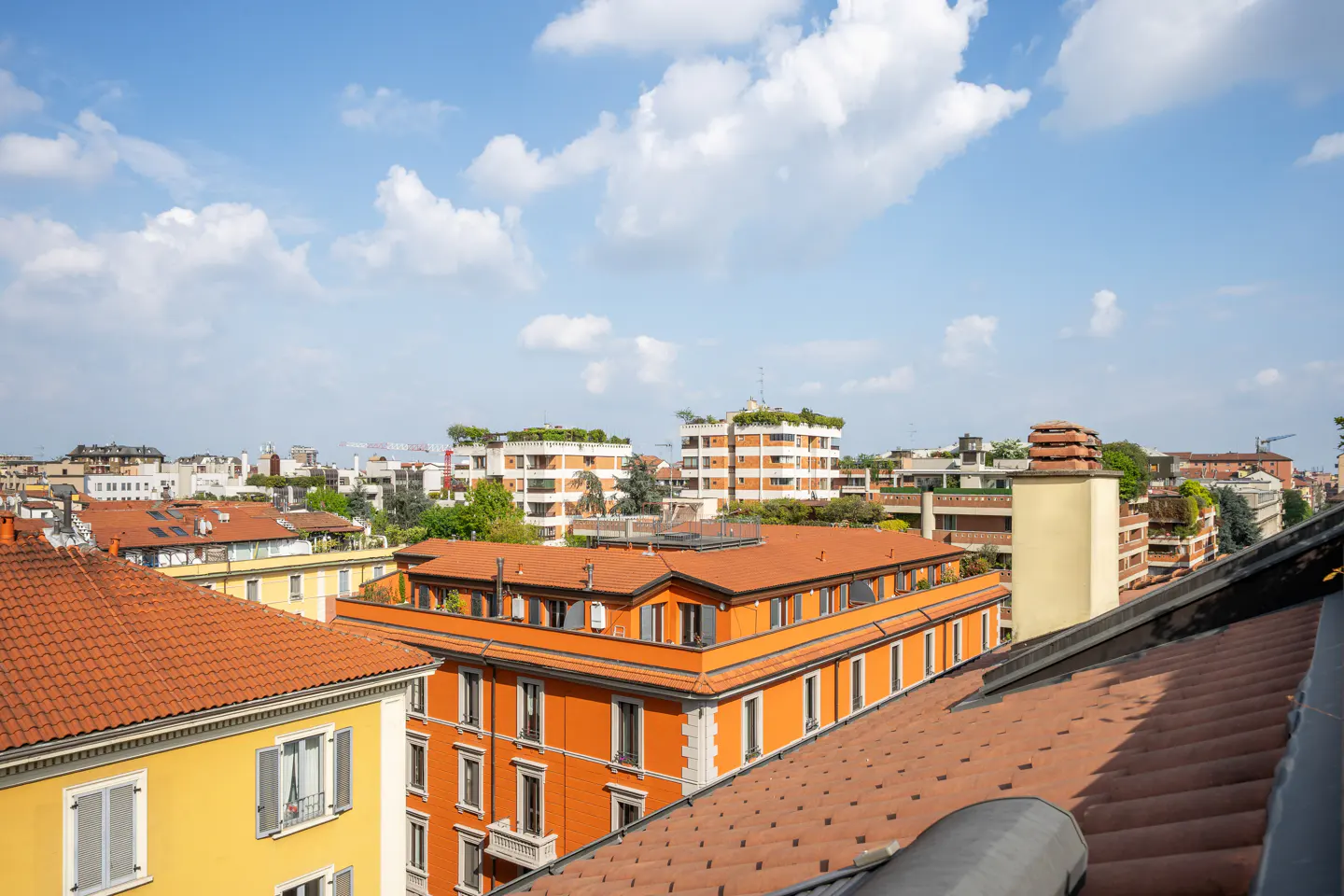 Cityscape view with orange and yellow buildings under a blue sky with white clouds. Rooftops are visible in the foreground.