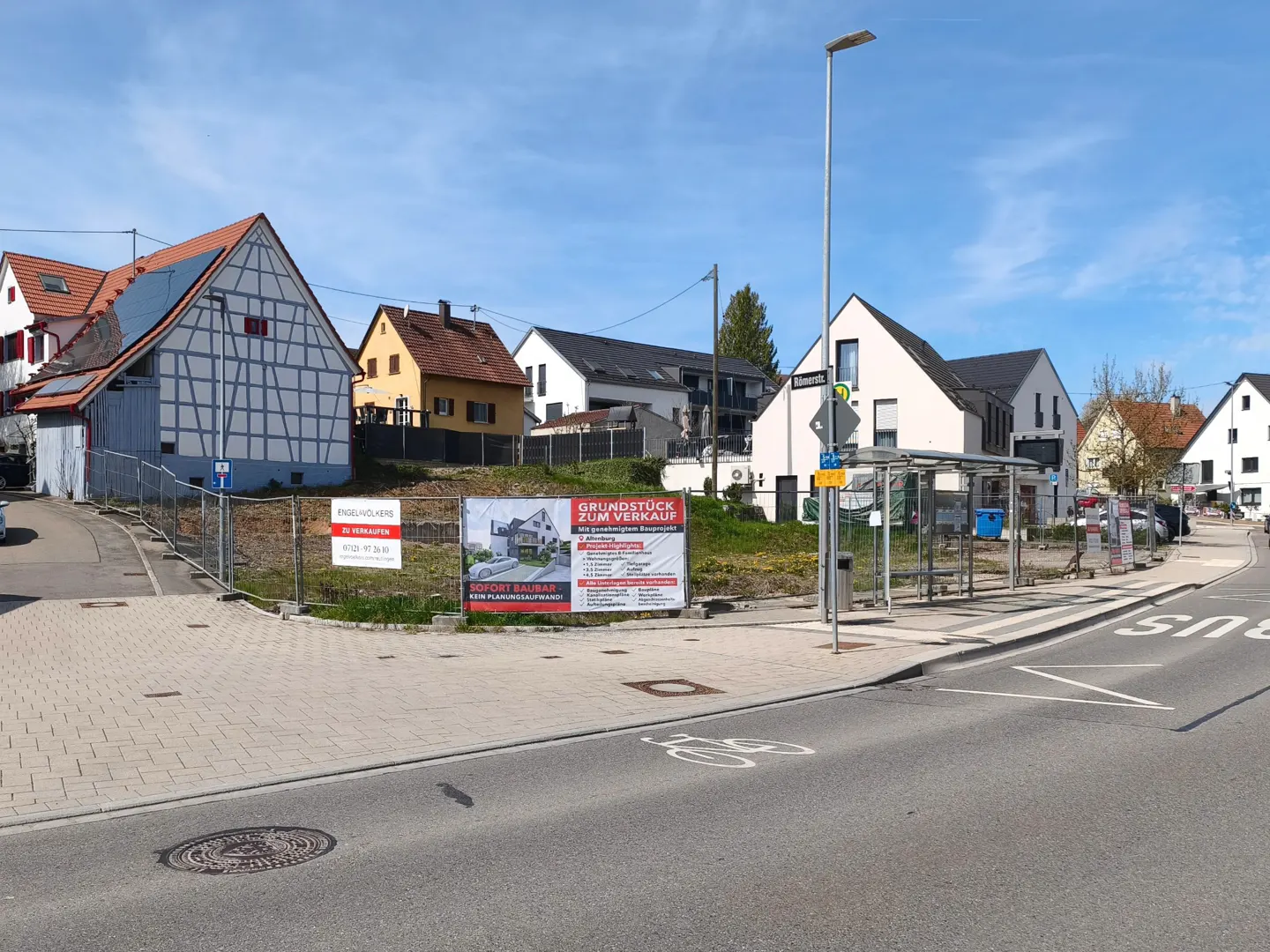 A vacant lot with "For Sale" signs sits near a bus stop on a street lined with houses under a blue sky.