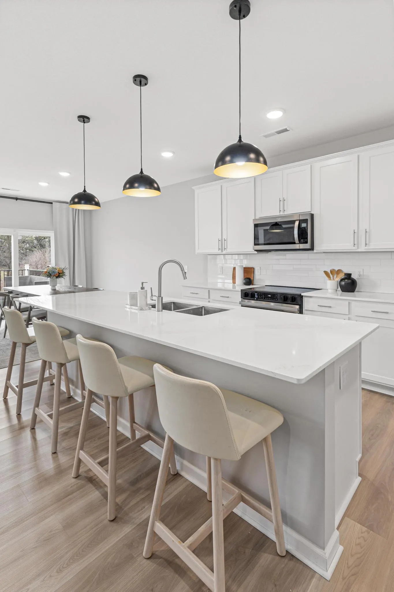 Bright, modern kitchen with white cabinets, quartz countertops, and wood floors. Black pendant lights hang above a large island with four beige stools.