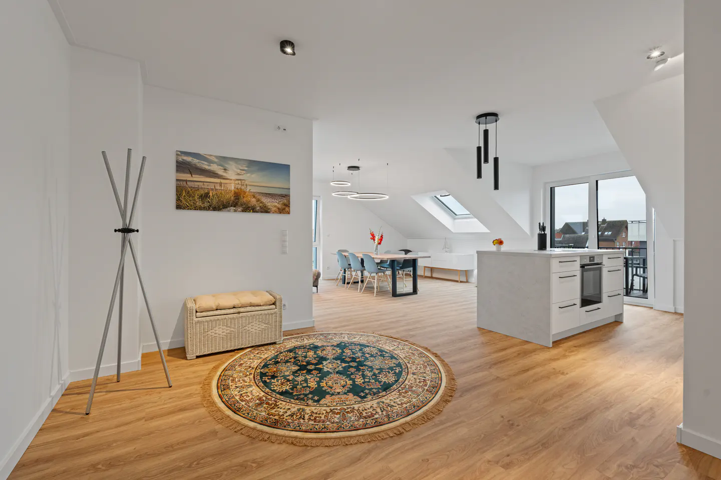 Bright, modern apartment interior with wood floors, white walls, and a round rug. A coat rack and bench sit near a beach scene art piece. The kitchen and dining area are visible in the background.