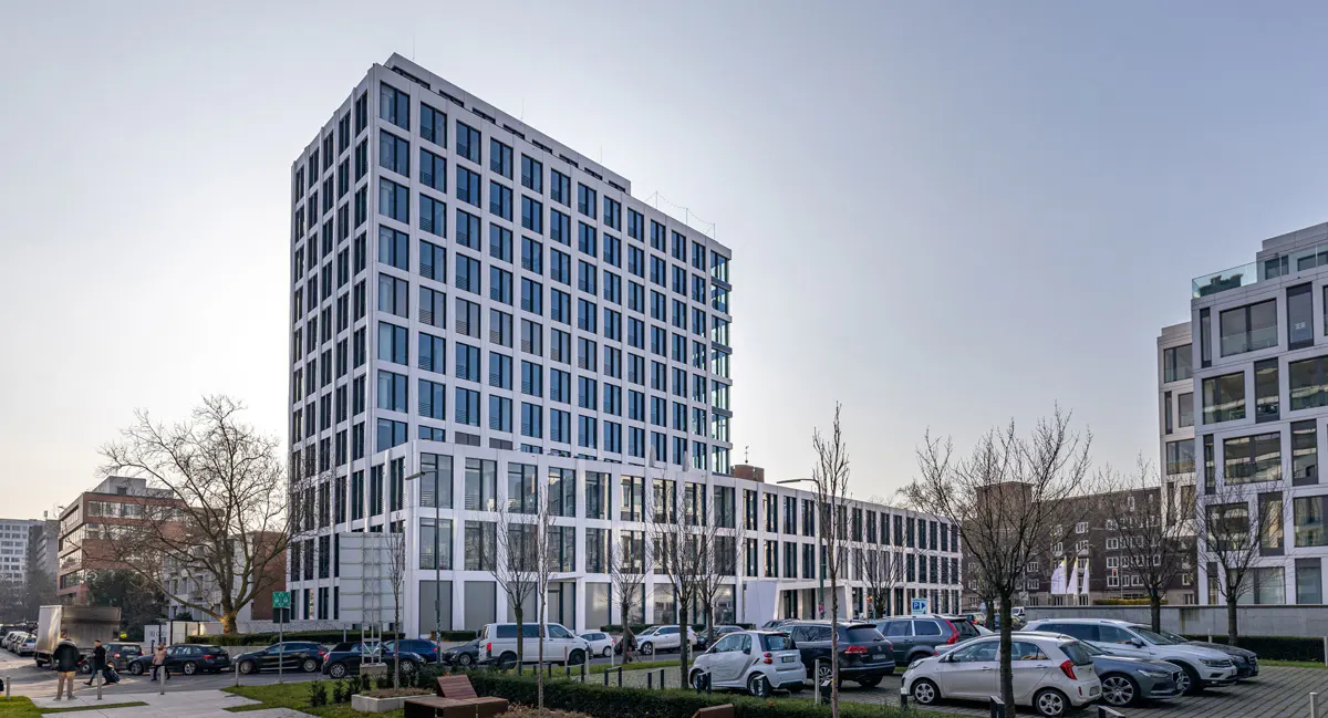 Modern white office building with many windows, parking lot in front, and trees.
