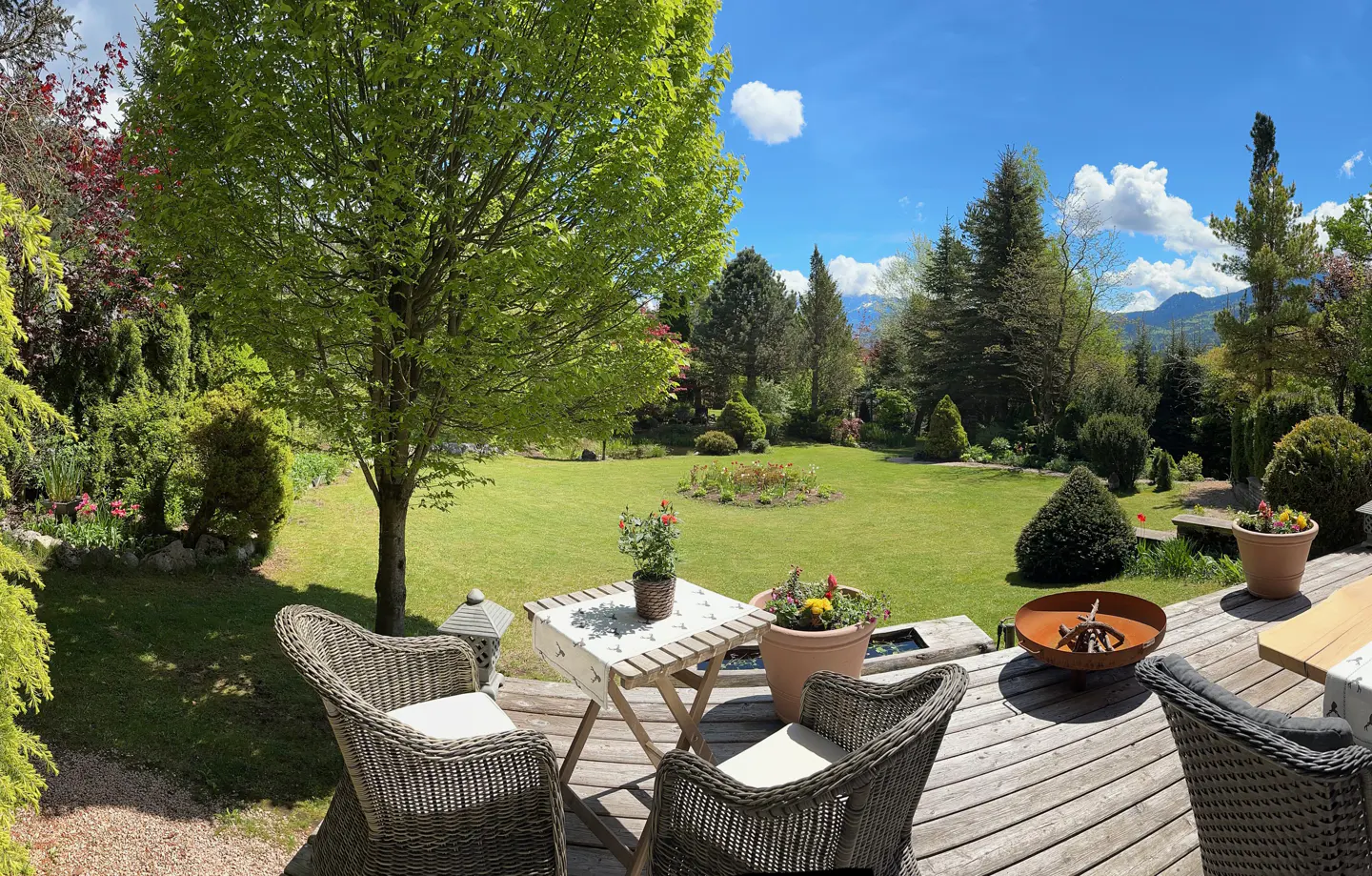 Wooden deck with wicker chairs and a small table overlooks a lush green lawn and garden with trees and mountains in the distance under a blue sky.