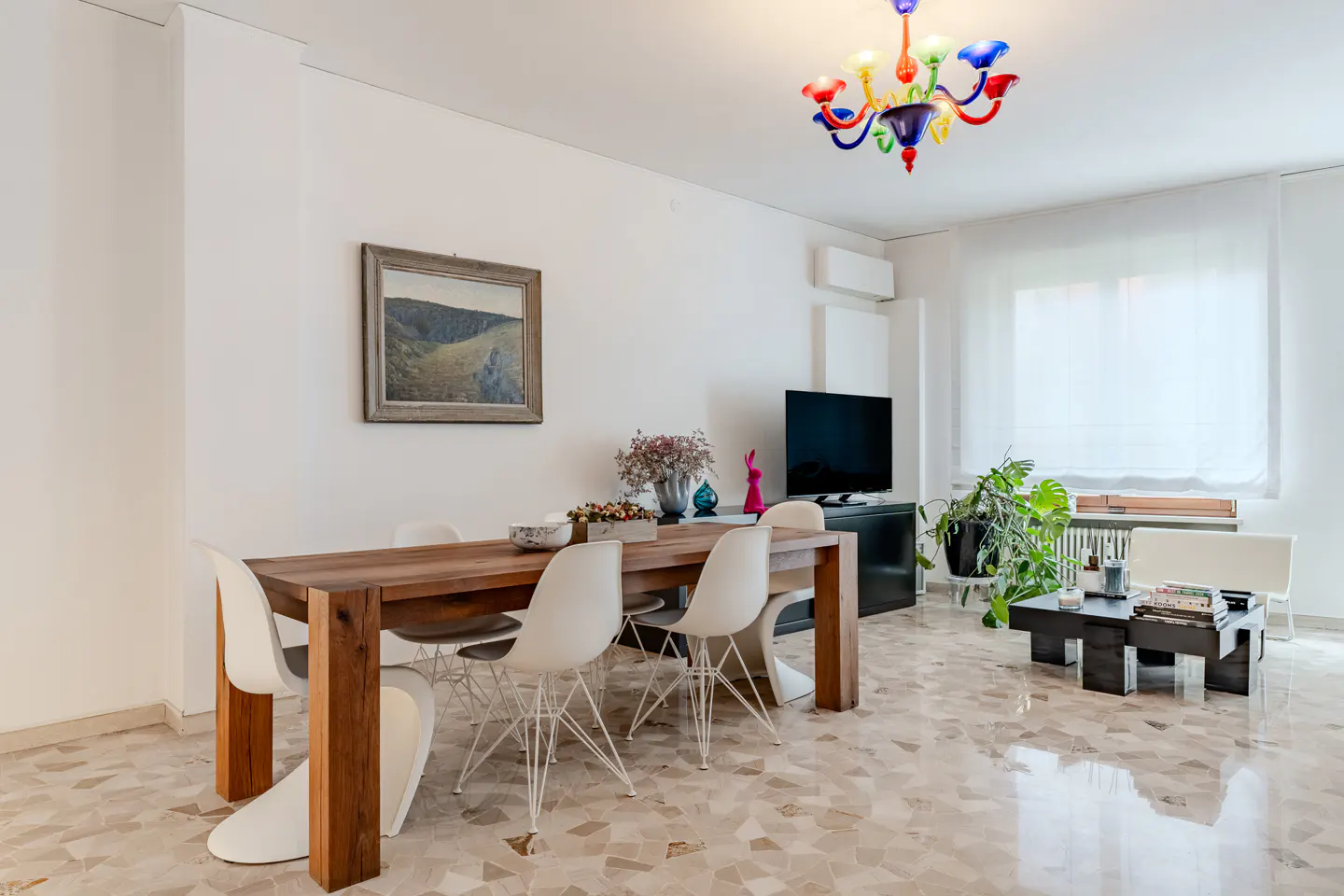 Bright dining room with a wooden table, white chairs, and a colorful chandelier. A painting hangs on the white wall. A TV and plants are in the background.