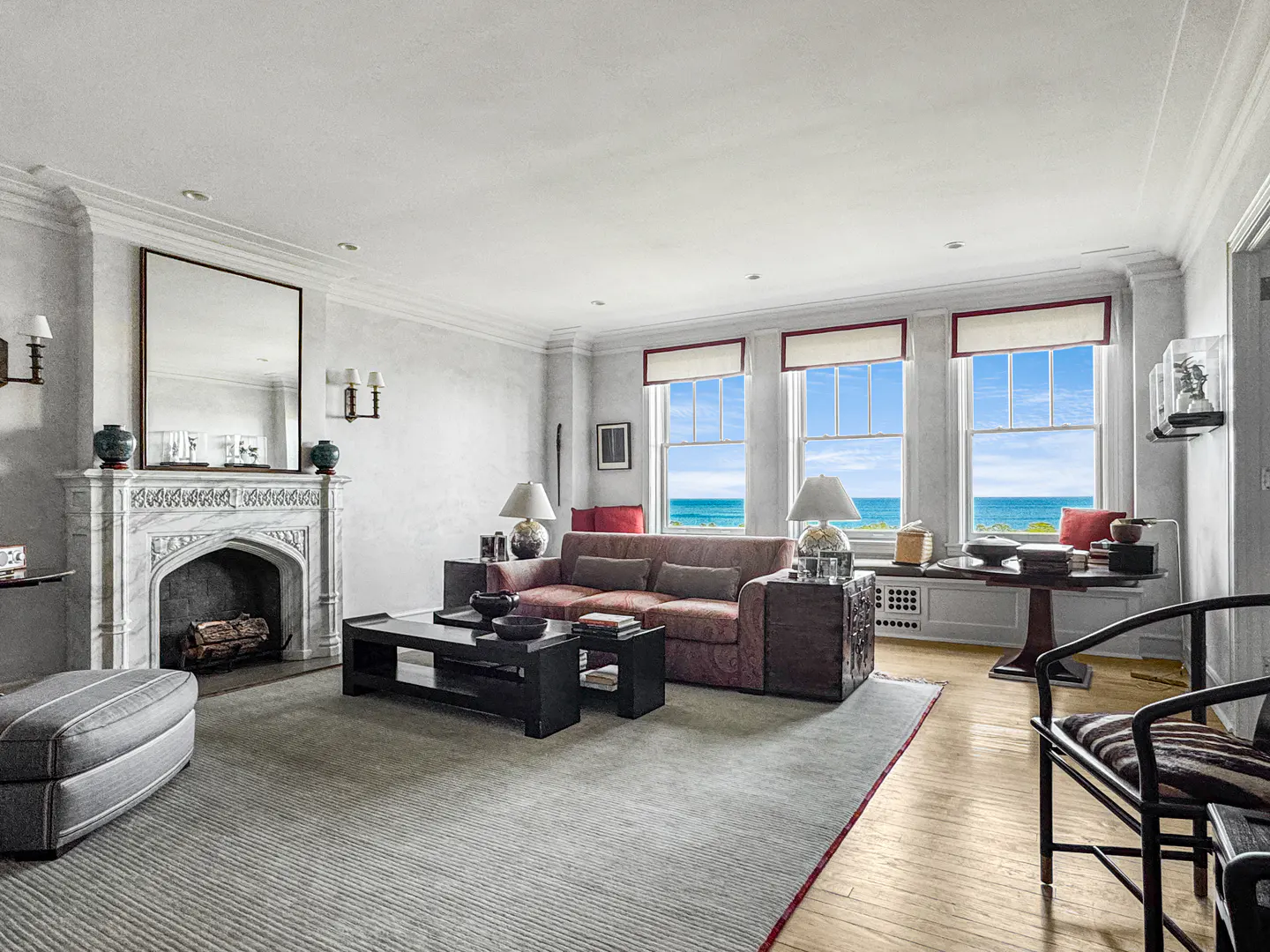Elegant living room with a marble fireplace, red sofa, and large windows offering a scenic ocean view.