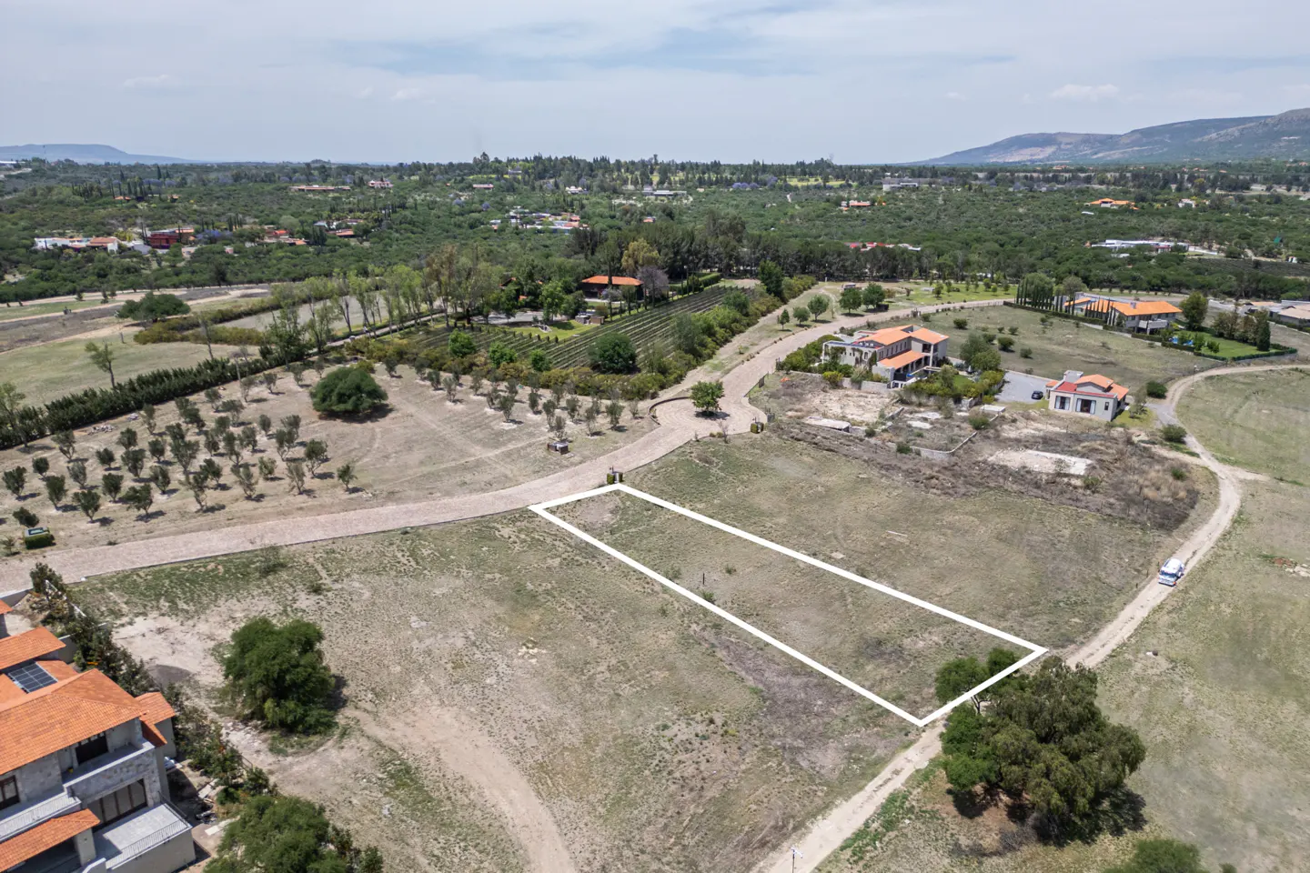 Aerial view of a vacant lot outlined in white, surrounded by trees, houses, and distant mountains.
