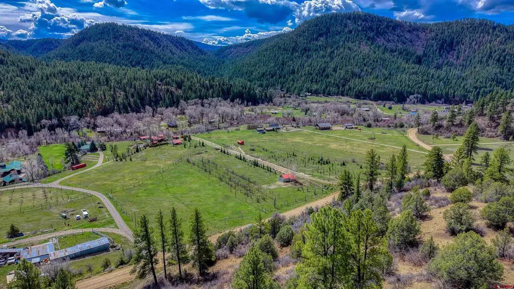 Aerial view of a green valley with scattered houses, surrounded by mountains and a blue sky with clouds.